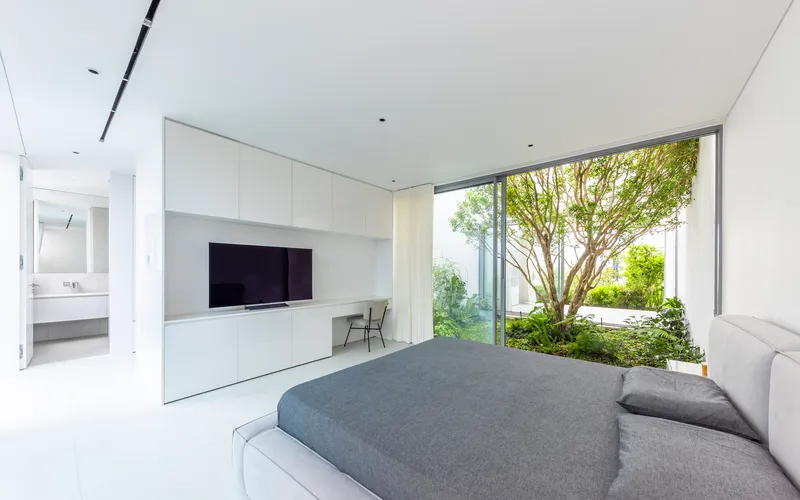 White bedroom with floor-to-ceiling glazing opening onto a courtyard garden with a mature tree