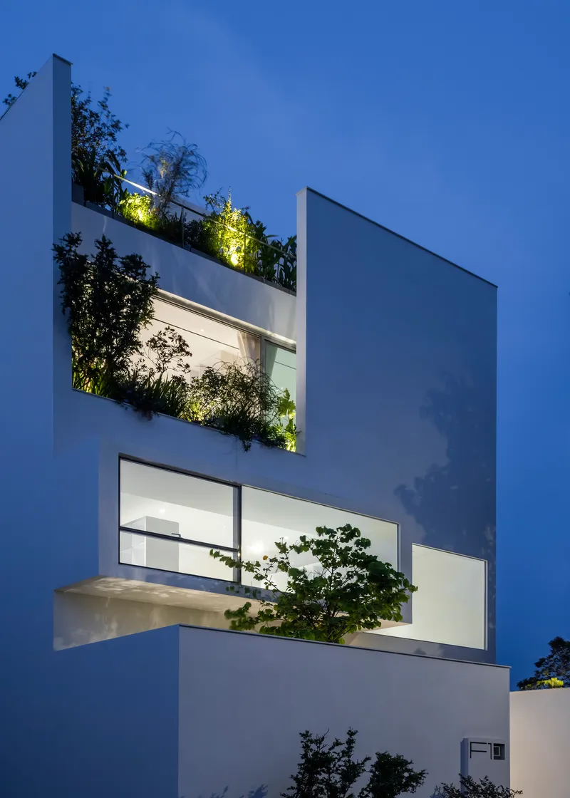 White facade with recessed planted terraces and illuminated greenery against a darkening evening sky