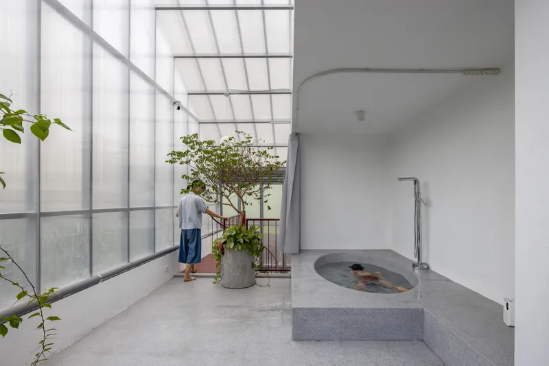 Interior bath terrace with raised concrete soaking tub and person standing near potted tree by translucent window wall