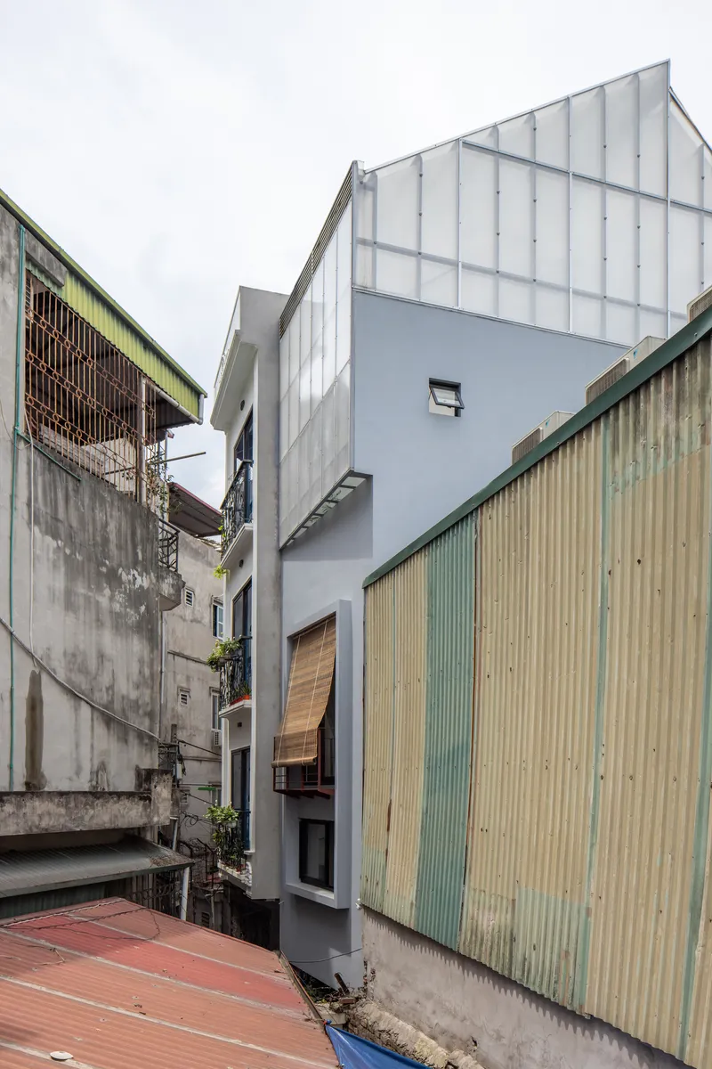 Narrow street facade with vertical translucent polycarbonate cladding flanked by weathered corrugated metal neighbors under cloudy sky