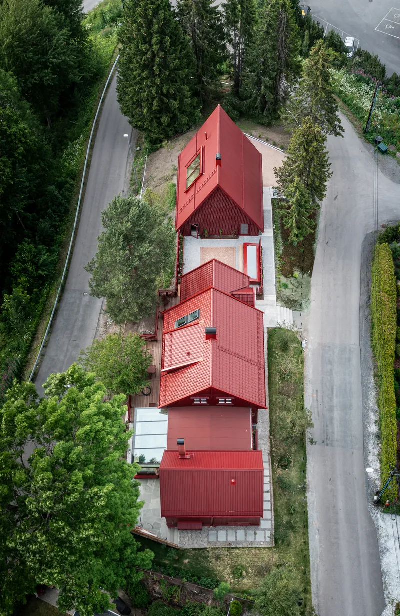Aerial view of four interlocking gabled volumes with red corrugated roofing nestled among conifers and asphalt driveways