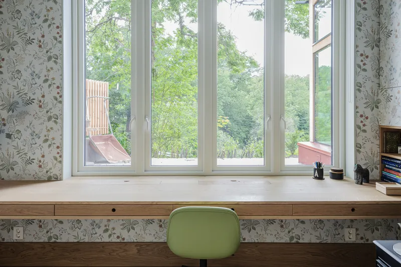 Built-in timber desk below a wide window bay with floral wallpaper and green chair