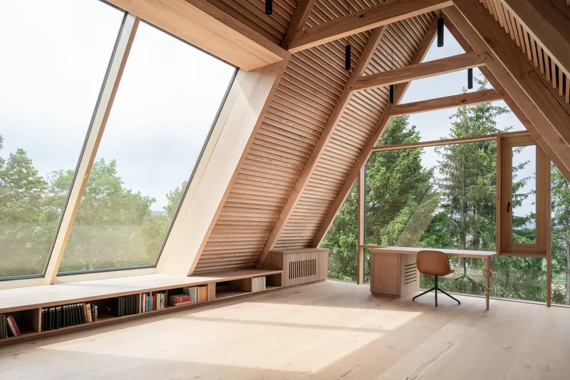 Sloped attic workspace with exposed timber rafters and angled glazing framing tree views