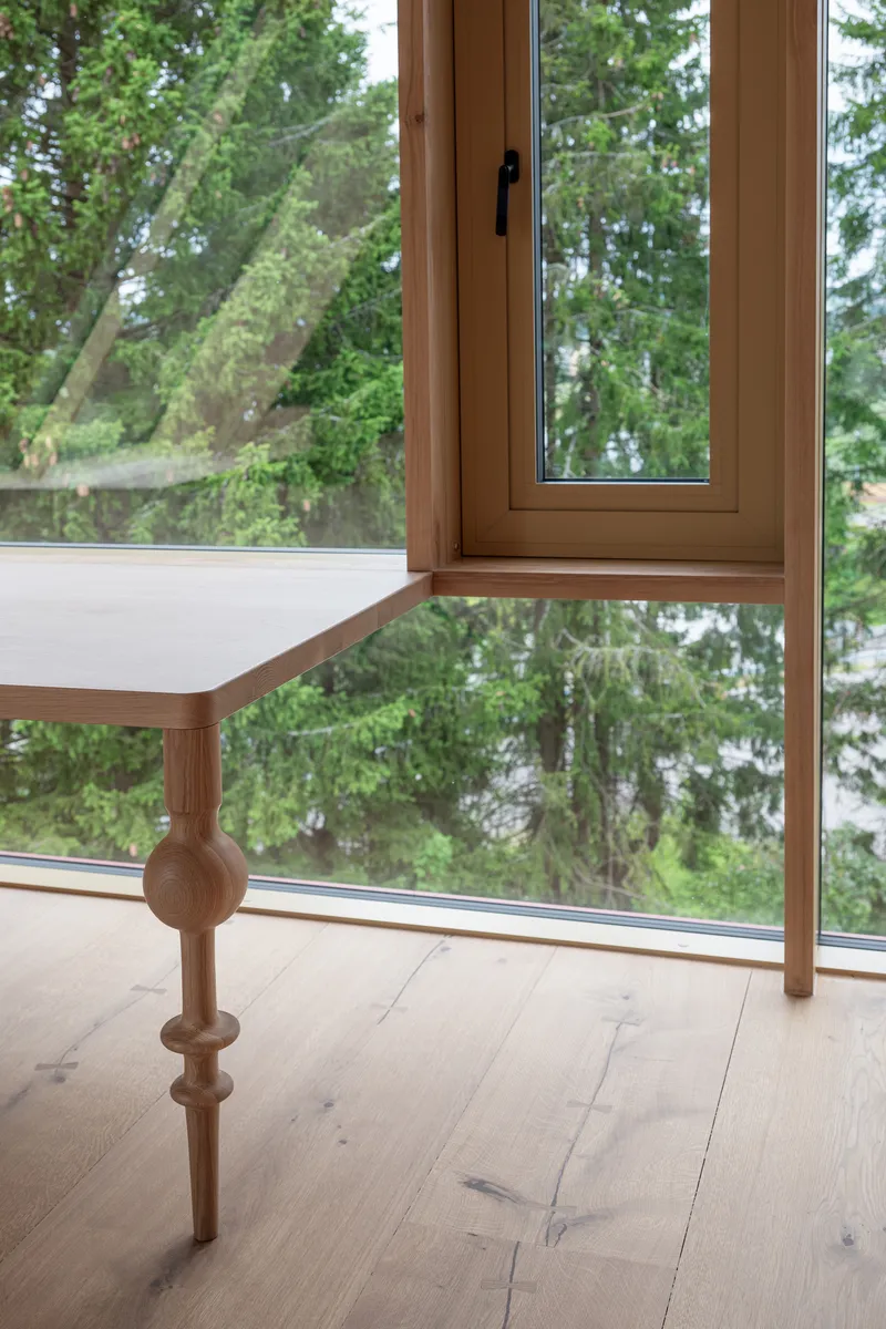 Turned timber balustrade detail with spherical joint beside floor-to-ceiling window overlooking forest