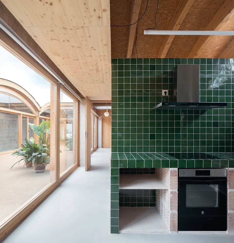 Dark green tiled kitchen volume with built-in oven beneath exposed timber ceiling and arched windows