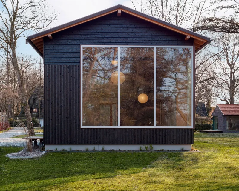 Charred timber gable facade with large tri-panel window reflecting bare trees in late afternoon light