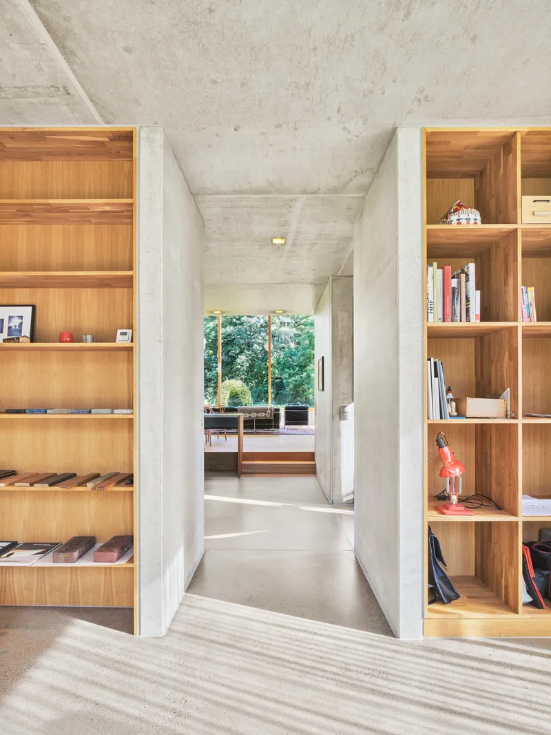 Interior hallway with exposed concrete ceiling flanked by built-in timber bookshelves and garden view beyond