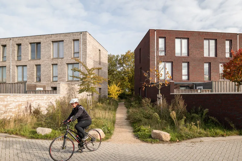 Cyclist passing between brick rowhouses separated by a gravel path and naturalized planting