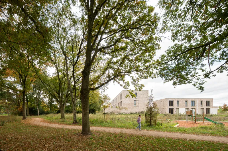 View of brick housing blocks across a grassy field with mature trees and a child on playground equipment