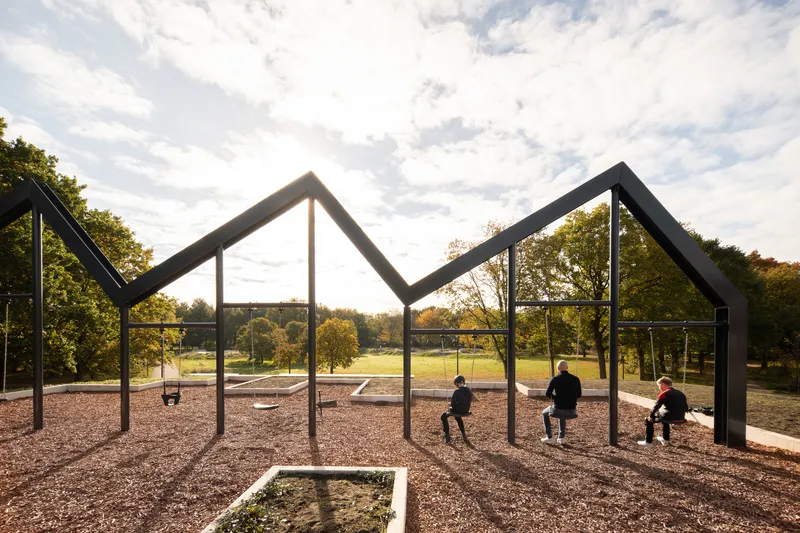 Steel frame house-shaped pavilion with three children seated on swings under a partly cloudy sky
