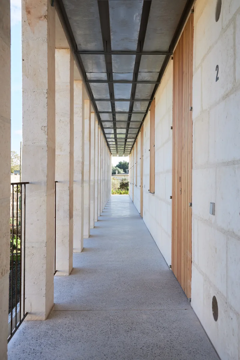 Long exterior corridor with stone columns, steel and glass ceiling panels, and timber doorways along the wall
