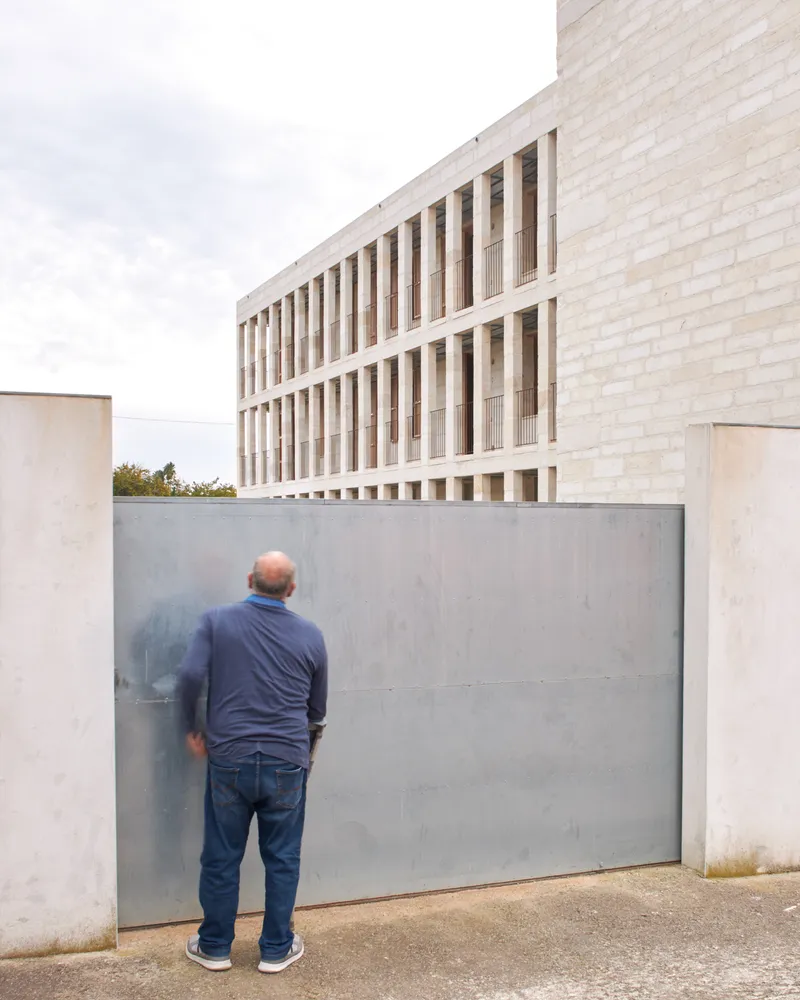 Figure standing at a steel gate below the gridded stone facade in soft light