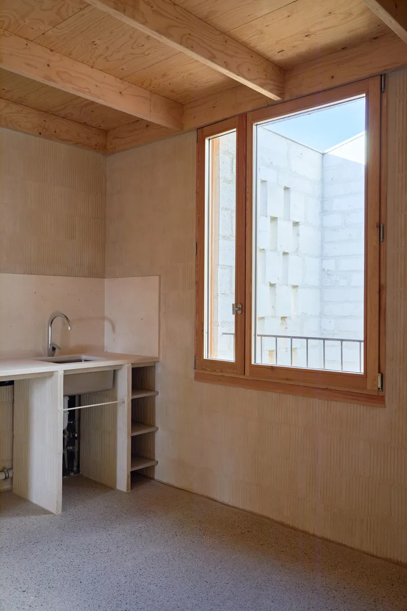 Small kitchen counter with open shelving beneath timber-framed casement window overlooking a neighboring white facade