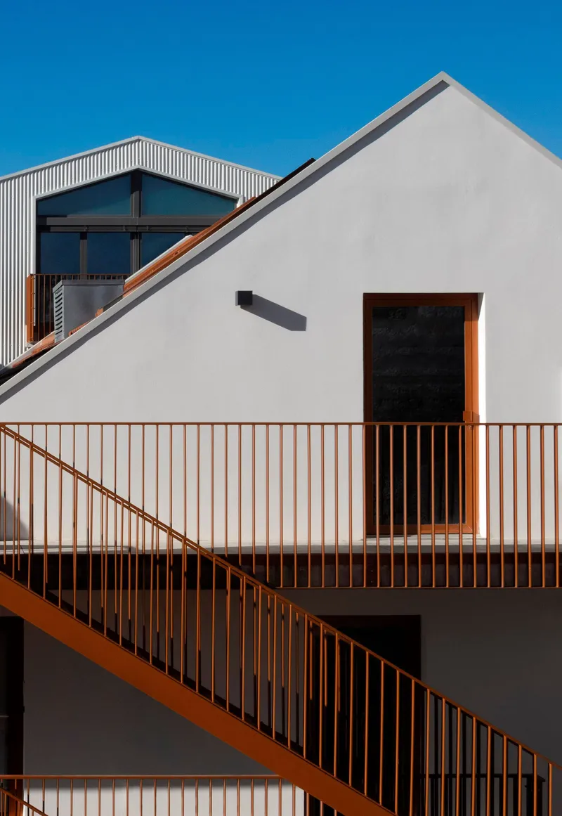 White rendered facade with timber-railed balcony and diagonal orange steel staircase under clear blue sky