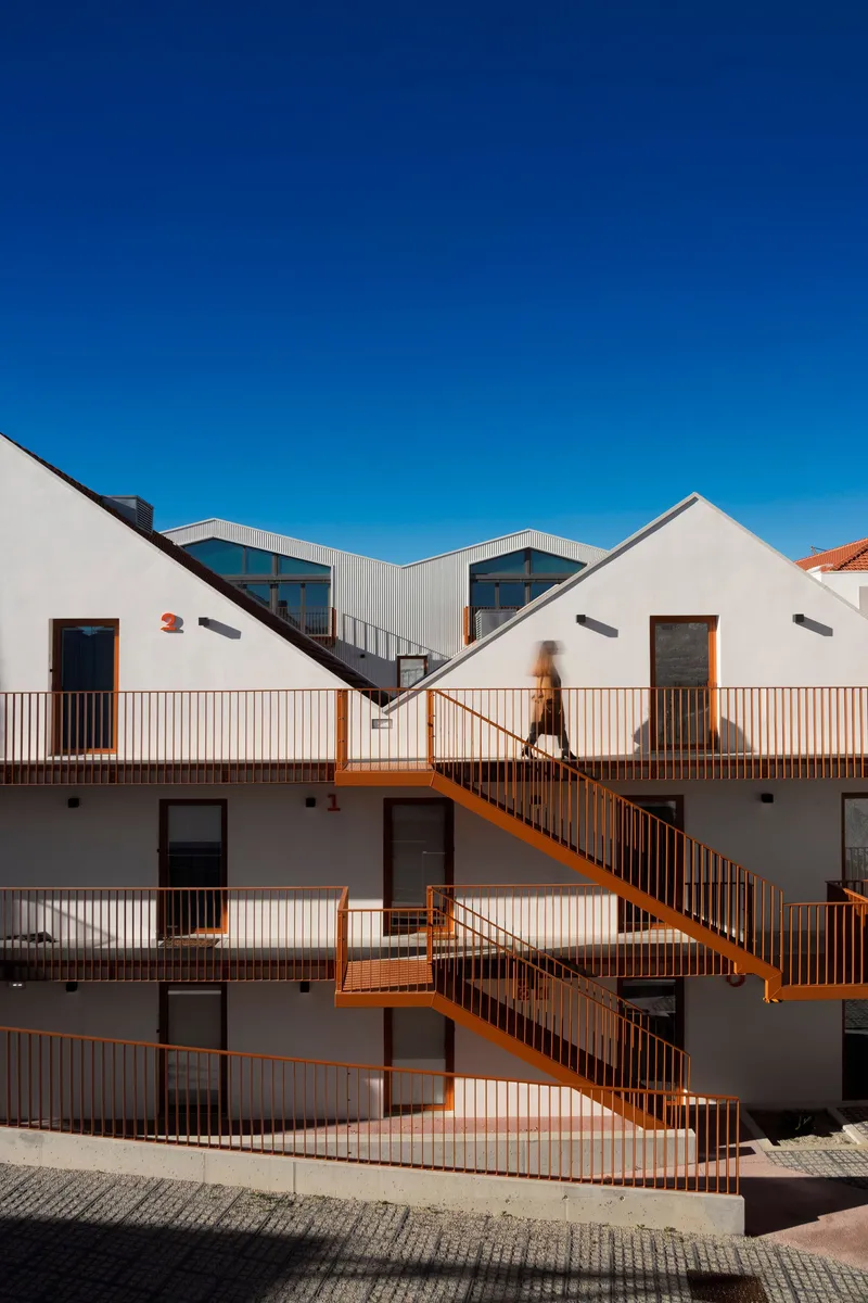 Courtyard facade with zigzagging orange steel stairs connecting white volumes and timber balconies as figure ascends