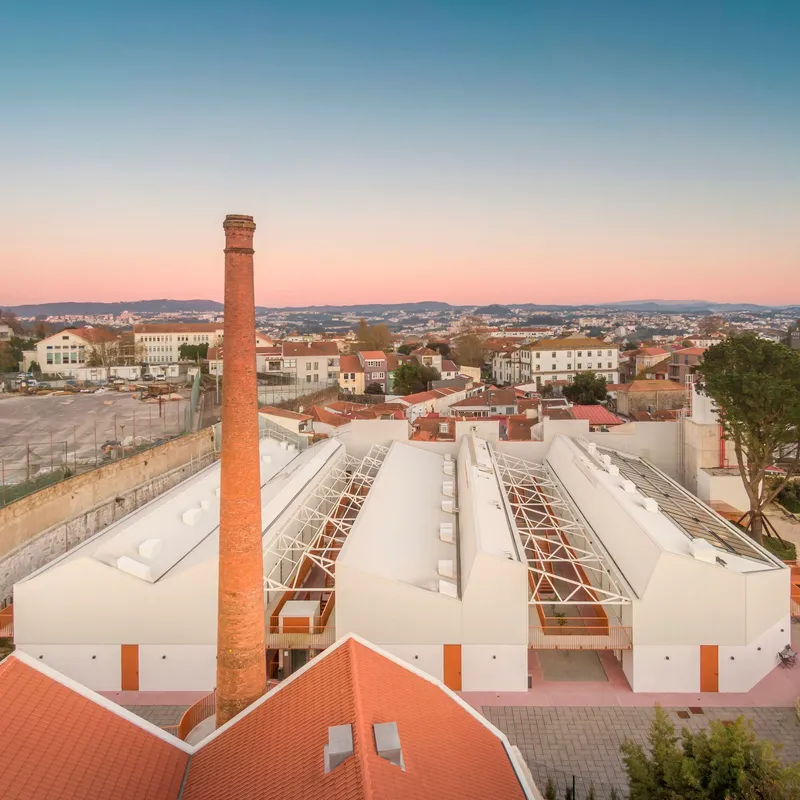 Aerial view of parallel sawtooth roofs with glazed sections and a tall brick chimney at dusk