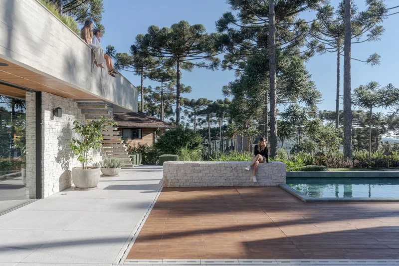 Pool terrace with person walking past timber deck panels and planted concrete wall under araucaria trees