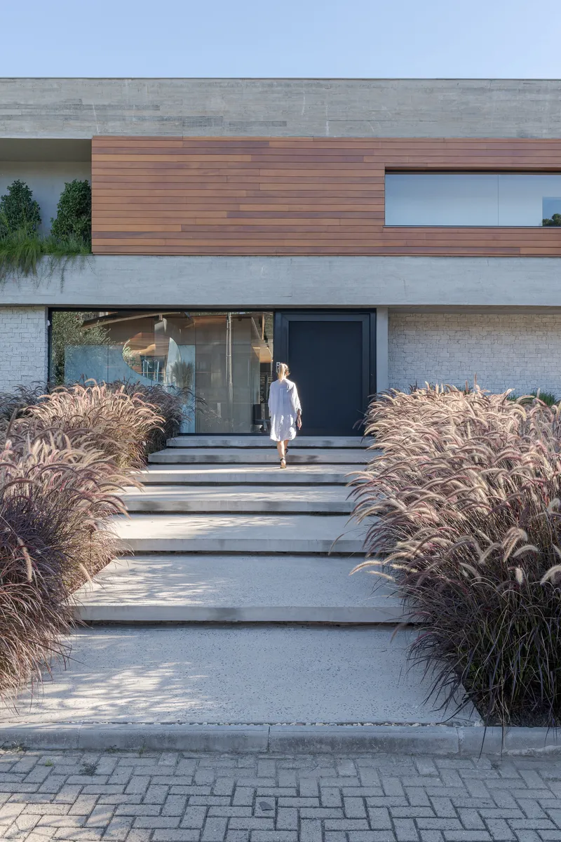 Entry steps flanked by dried ornamental grasses with person approaching dark doorway beneath timber and concrete facade