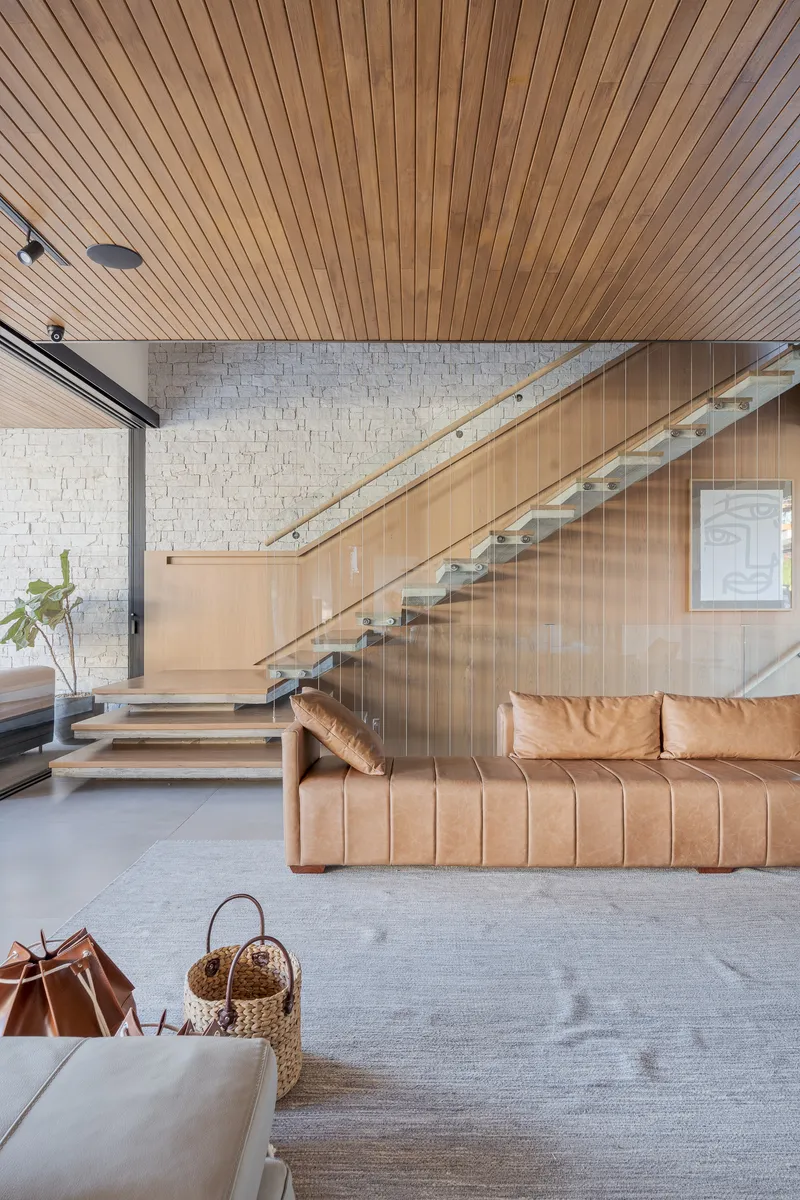 Floating timber and glass staircase ascending beneath a slatted wood ceiling and white brick wall