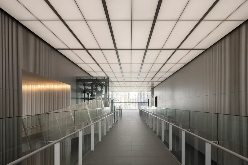 Interior bridge with glass railings under a gridded translucent ceiling framed by ribbed metal walls