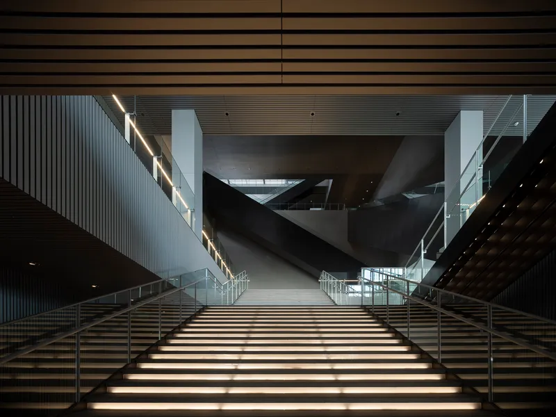 Wide staircase with integrated lighting rising toward angular volumes suspended in the atrium