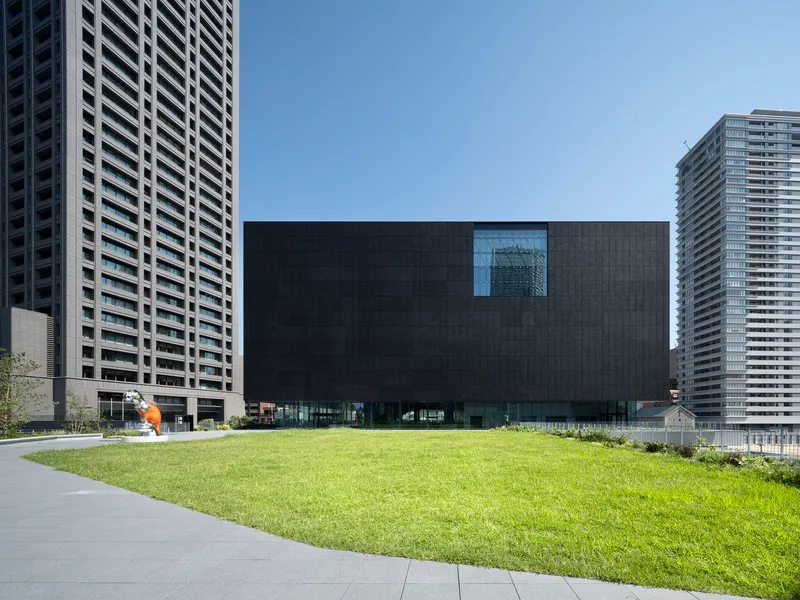 Dark textured facade with single recessed window facing a lawn and residential towers beyond