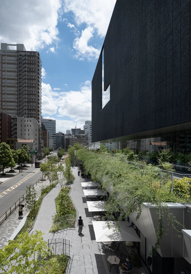 Linear planted promenade with trees and pedestrians running alongside the elevated dark volume
