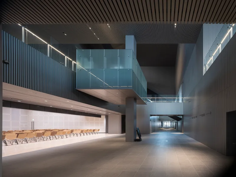 Interior lobby with slatted ceiling, suspended glass bridge and rows of wooden chairs in side gallery
