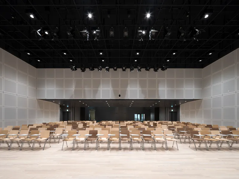 Performance hall with exposed rigging, white panel walls and wooden seating on pale flooring