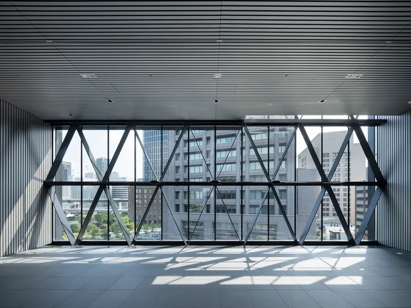 Floor-to-ceiling triangulated steel bracing behind glass with sunlight creating geometric shadow patterns on tile flooring