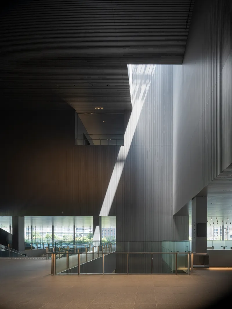 Double-height atrium with tilted ribbed wall catching sharp daylight from an overhead skylight