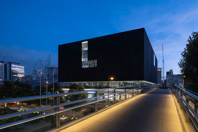 Black cubic volume elevated on slender columns viewed from pedestrian bridge at dusk