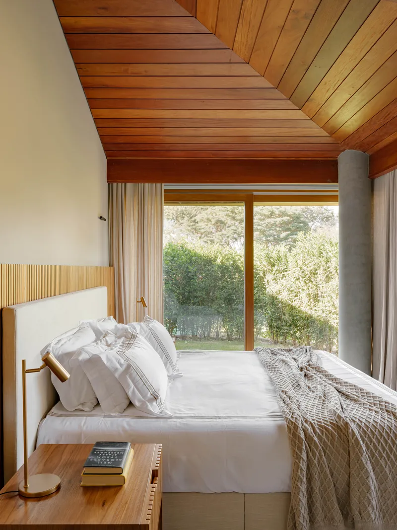 Bedroom with timber slatted ceiling, timber-framed glazing overlooking garden and cylindrical concrete flue column