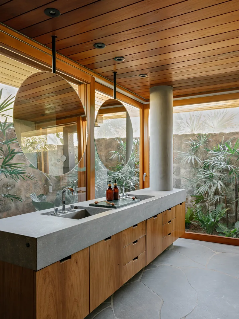 Bathroom vanity with concrete countertop, timber cabinetry, oval mirrors and planted courtyard beyond glass wall