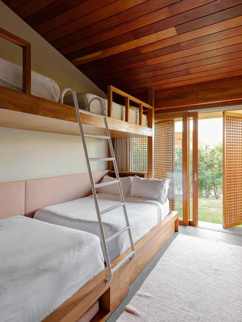Bunk bedroom with timber-framed beds, metal ladder, sloped timber ceiling and screened sliding doors to garden