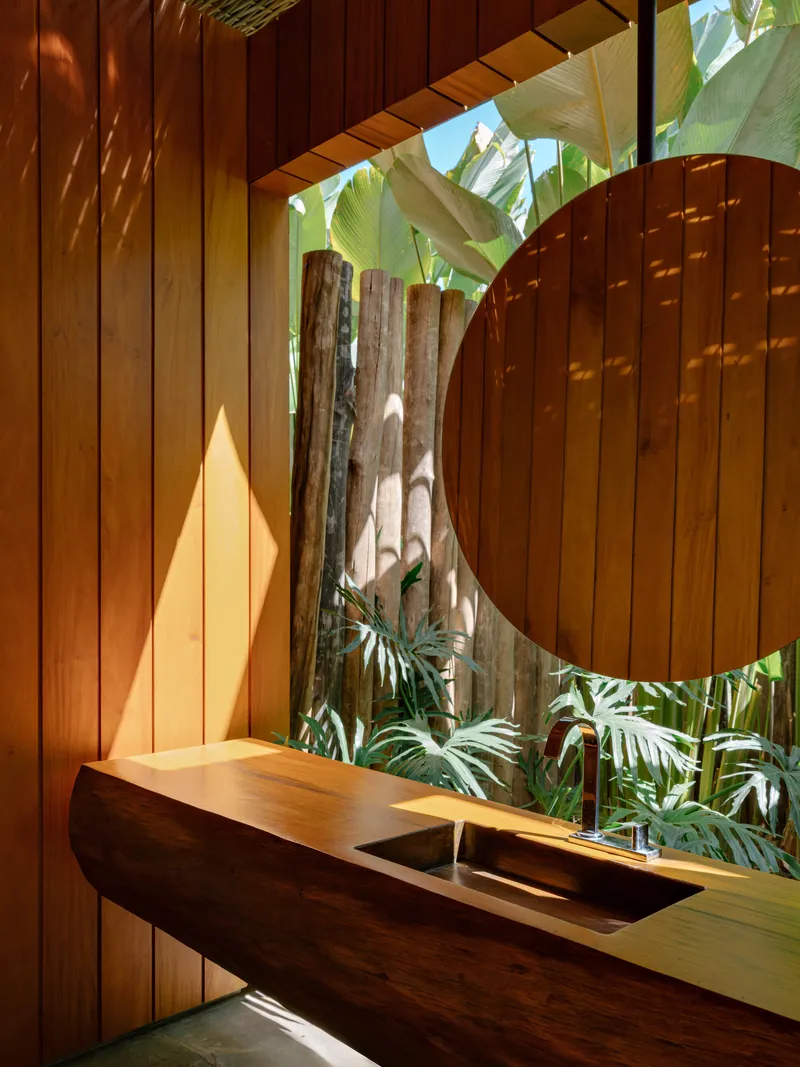 Timber vanity counter beneath a circular cutout in vertical wood slat wall with tropical plants beyond