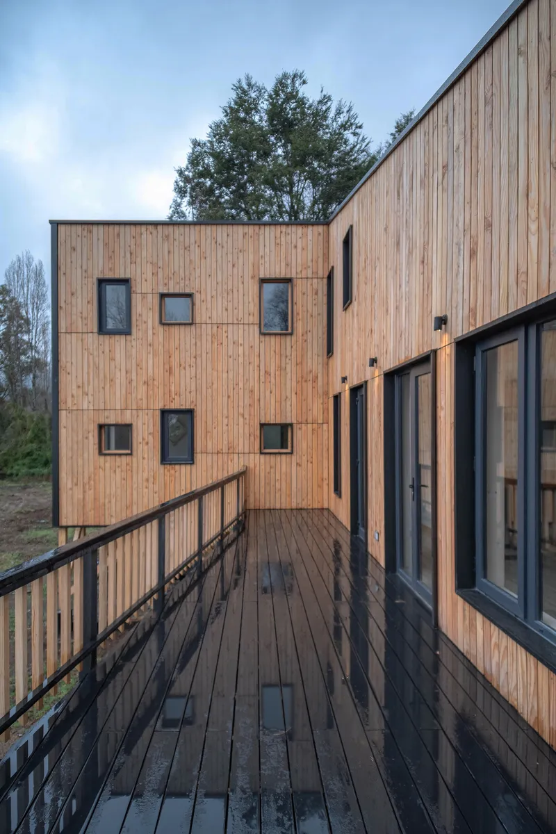 Timber deck connecting vertical wood-clad facades with black-framed windows under evening sky