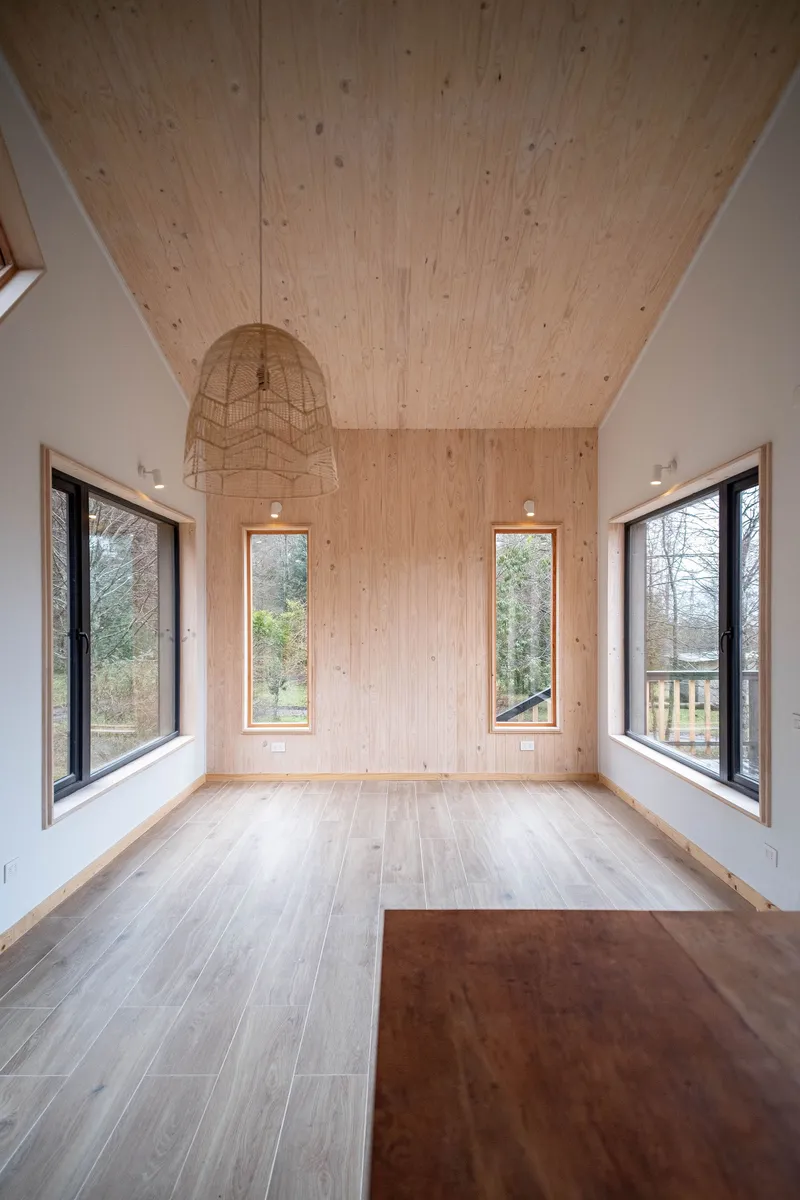 Interior room with exposed plywood ceiling panels and wire pendant light between tall black-framed windows