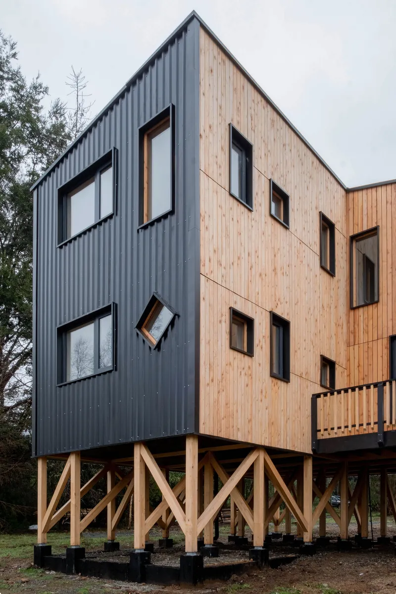 Split facade showing half in dark corrugated metal and half in vertical timber siding on timber piles