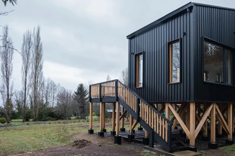 Exterior stair with timber railing leading to elevated metal-clad volume on diagonal braced posts