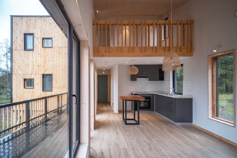 Double-height interior with timber slat mezzanine railing above a galley kitchen with sliding glass doors to the deck