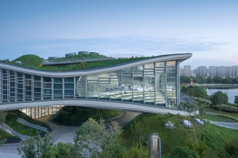 Glass curtain wall and cantilevered roof edge at dusk with planted slopes descending to the ground level