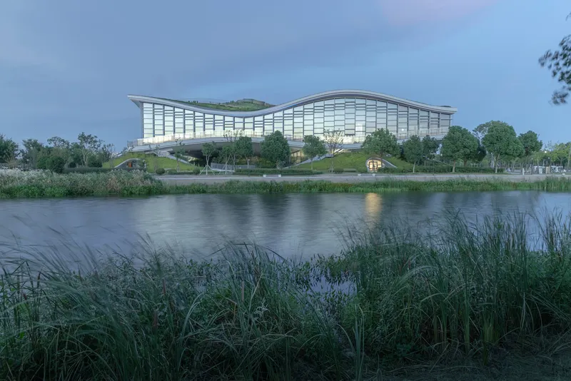 Glass and steel facade with planted roof seen across the water at twilight with reeds