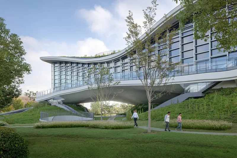 Cantilevered glass-enclosed bridge structure spanning landscaped slopes with young trees and visitors walking beneath