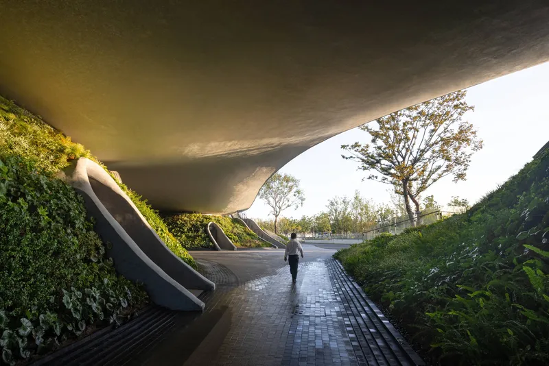 Pedestrian tunnel beneath a sweeping concrete deck with planted embankments and paved walkway at dusk