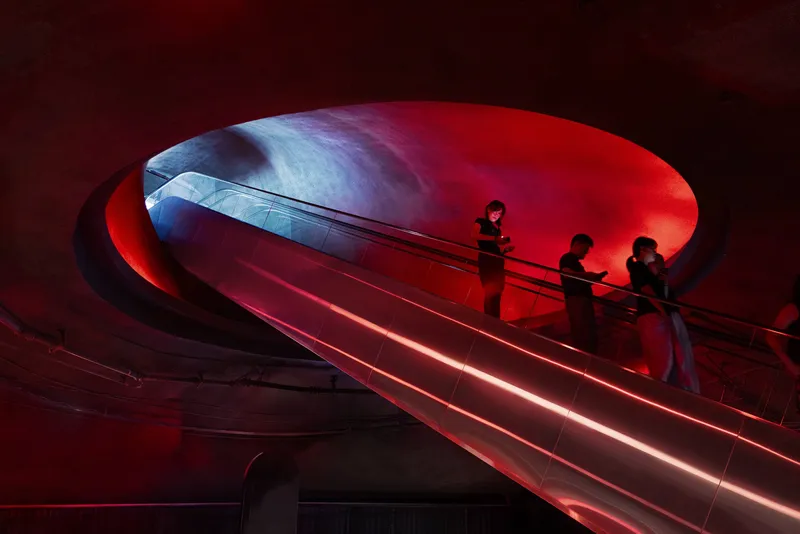 Helical escalator rising through an elliptical void bathed in red and blue gradient lighting