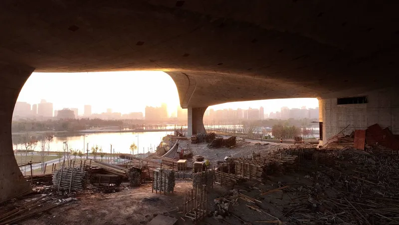 Arched openings in raw concrete shell framing a waterfront skyline at sunrise during construction
