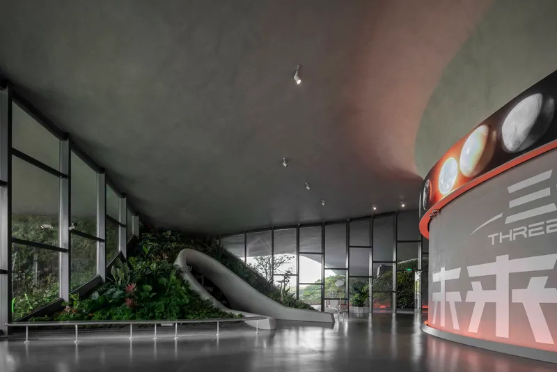 Interior space with floor-to-ceiling glazing, planted hillocks, and a tubular slide under concrete ceiling