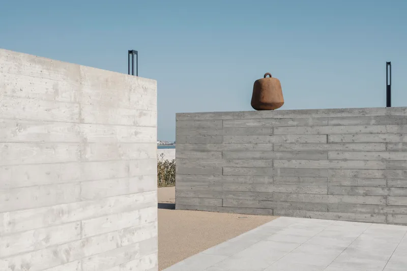 Courtyard framed by board-formed concrete walls with weathered bell suspended from upper parapet