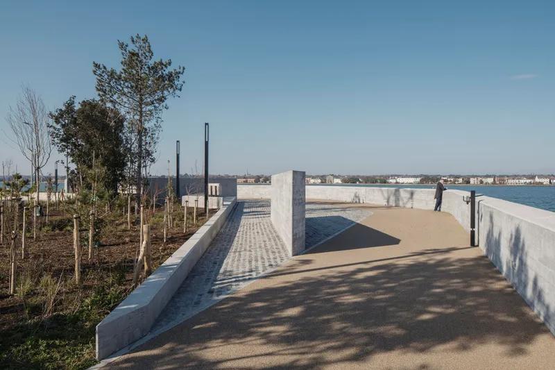 Curving pathway bordered by concrete wall and planted beds with young trees overlooking the bay